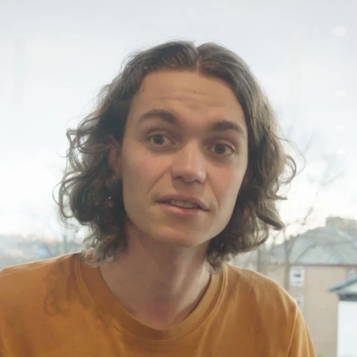 Person with curly hair wearing a mustard-colored shirt, blurred background.