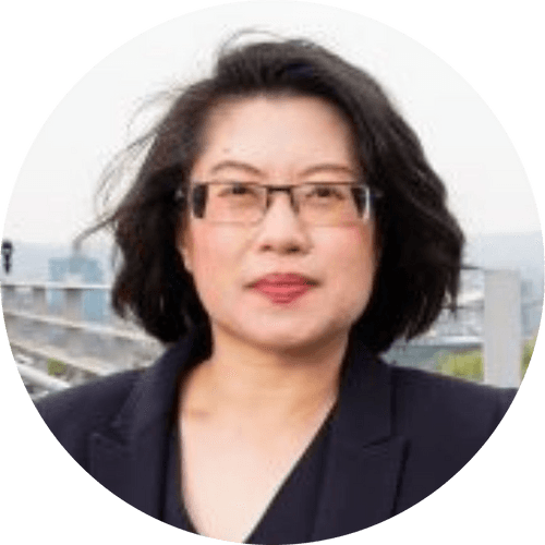 Person wearing glasses and a blazer, outdoors with blurred cityscape background.