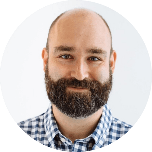 Man with a beard wearing a checkered shirt, smiling at the camera against a plain background.